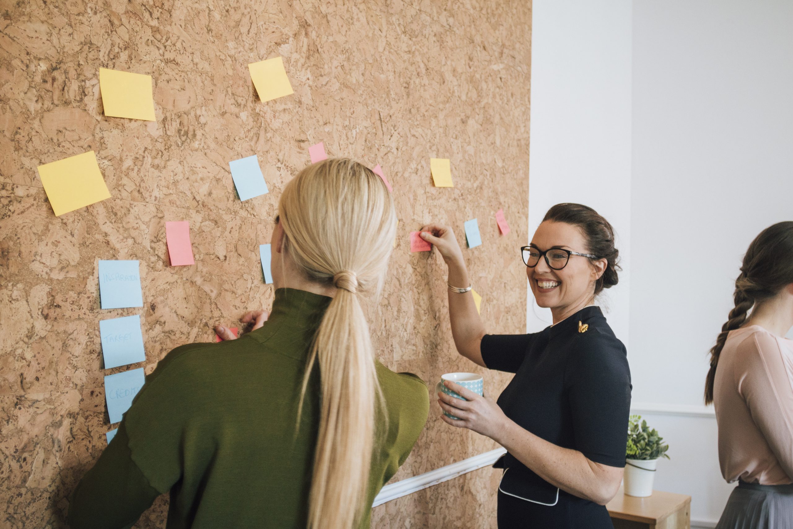Women Making Business Notes Two women are at work in an office. They are standing at a cork board and are having a discussion as they pin things up.